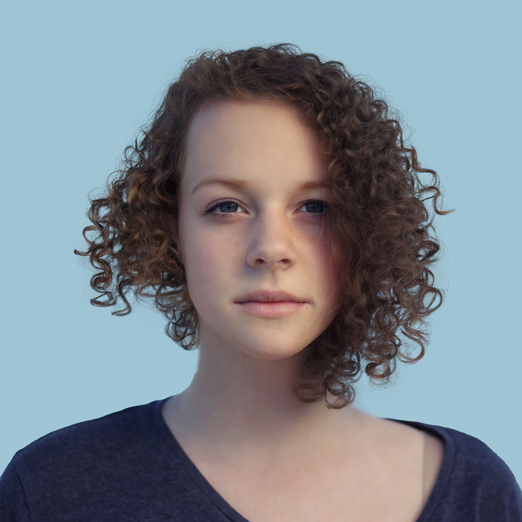 A young person with curly brown hair and a blue shirt stands against a plain light blue background, looking directly at the camera, exuding a calm and confident demeanor.