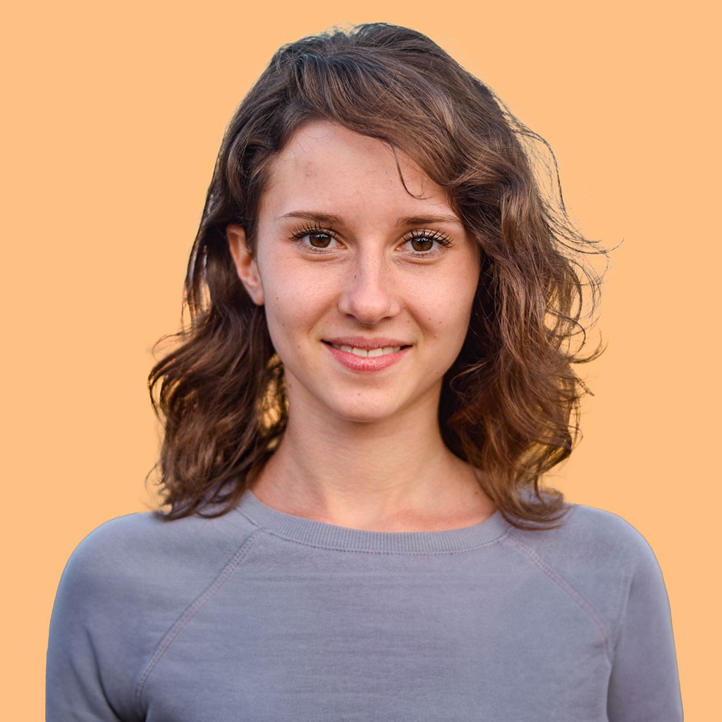 A woman with wavy, shoulder-length brown hair smiles while looking directly at the camera, standing against a solid orange background, wearing a casual gray sweater.